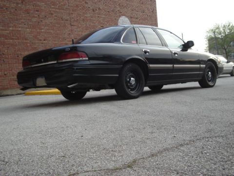 1992 Ford Crown Victoria - This black sedan is parked on a street with a brick building to its left and another vehicle in the background.