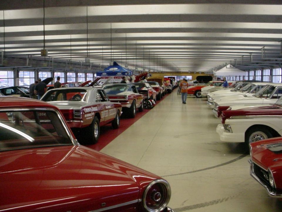 Indoor car show with rows of classic cars, some with hoods open. A few people are walking around
