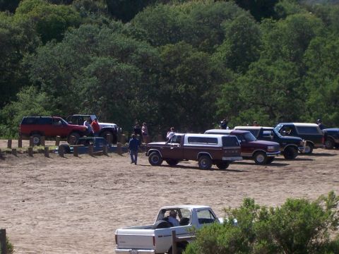1991 Ford Bronco - Several trucks and SUVs are parked on a dirt lot with trees in the background.