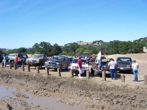 1991 Ford Bronco - A group of off-road vehicles are parked on a dirt track next to a muddy obstacle course.