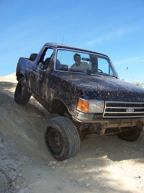1991 Ford Bronco - A driver navigates a muddy Ford Bronco up a steep, sandy incline under a clear blue sky.