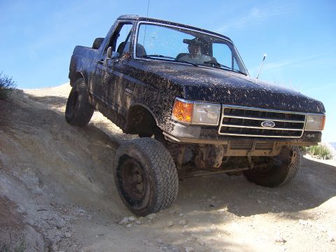 1991 Ford Bronco - A muddy black Ford Bronco navigates a steep dirt incline under a clear blue sky.