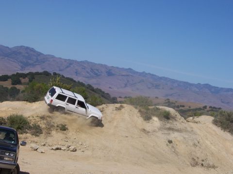 1991 Ford Bronco - A white Jeep Cherokee navigates a steep dirt trail with a dark pickup truck visible on the side.