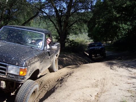 1991 Ford Bronco - Two trucks navigate a dirt trail, kicking up mud and dust under a canopy of trees.