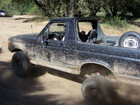 1991 Ford Bronco - A muddy Ford Bronco navigates a dirt path, kicking up dust.
