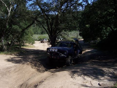 1991 Ford Bronco - Multiple off-road vehicles traverse a dusty trail surrounded by trees during an outdoor event.