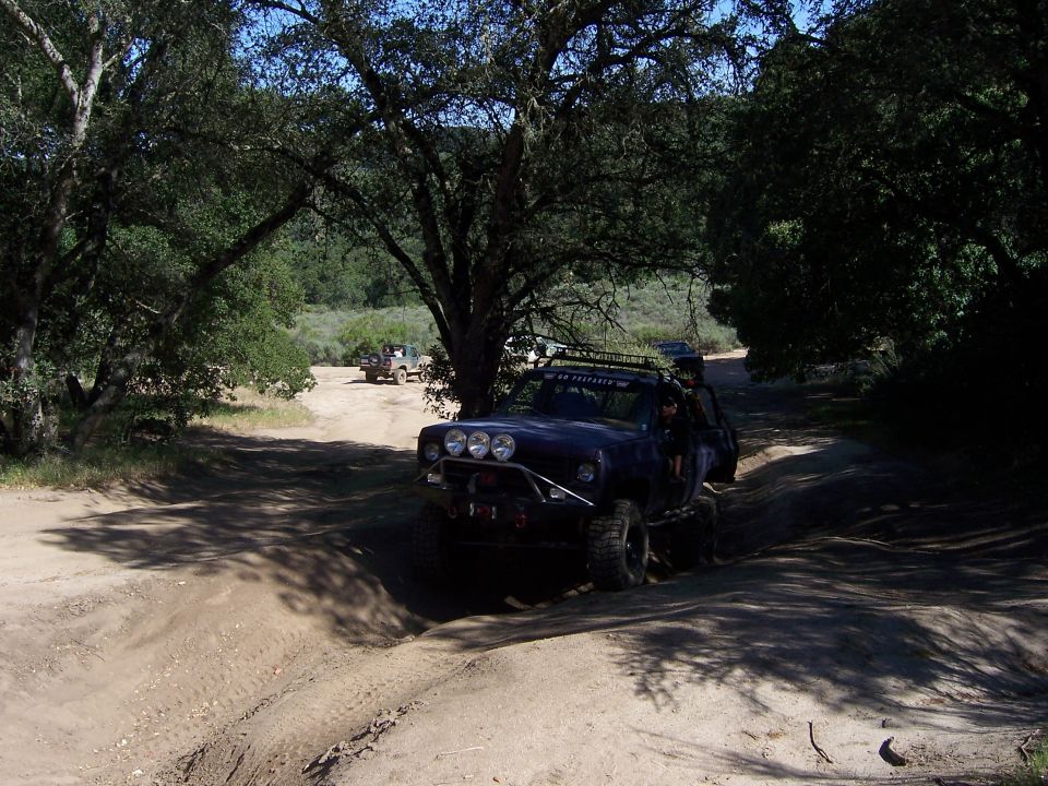 Off-road vehicles navigating a dirt trail under trees. One vehicle is in the foreground, modified for off-roading