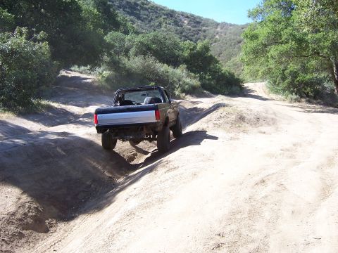 1991 Ford Bronco - A black Ford Bronco navigates a dusty, off-road trail surrounded by trees and hills.