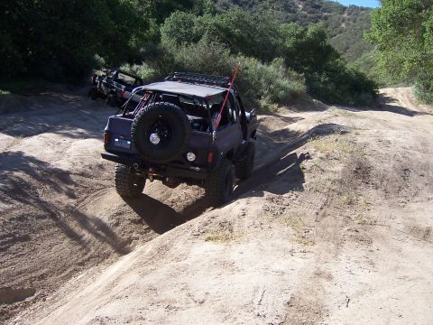 1991 Ford Bronco - Two off-road vehicles navigate a sandy, rutted trail on a sunny day with wooded hills in the background.