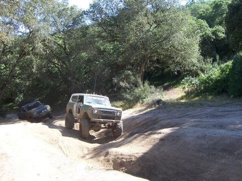 1991 Ford Bronco - Two off-road vehicles navigate a dusty trail surrounded by lush green trees on a sunny day.