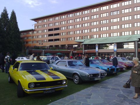 1991 Ford Crown Victoria - A bright yellow 1968 Chevrolet Camaro is parked on the grass in front of a modern building, with several other classic cars lined up behind…