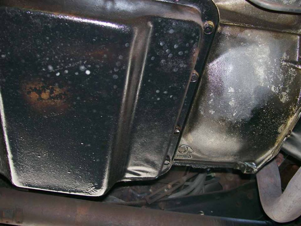Underside of a black steel oil pan meeting the bell housing, with rust and oil residue