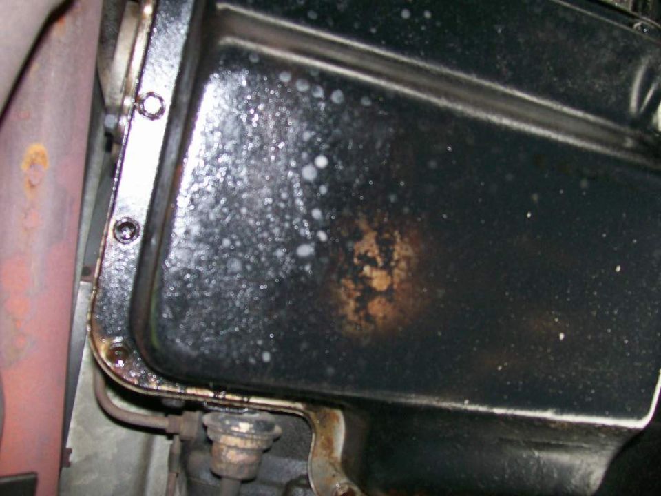 Black oil pan corner with rust bloom, white residue and flange bolt holes