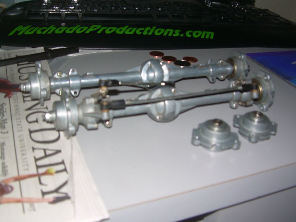 Two model vehicle axles and two differential covers sit on a surface with a newspaper and a keyboard in the background