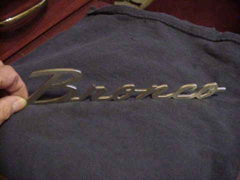 1993 Ford Bronco - A hand holds a chrome 'Bronco' emblem against a dark cloth background.