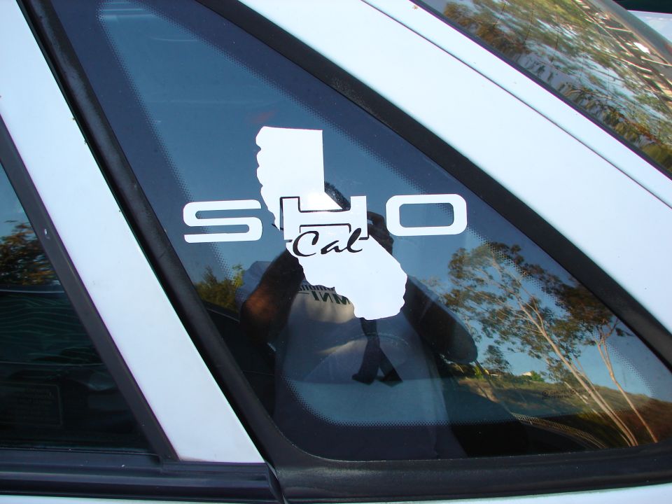 White SHO Cal sticker on a car's rear window, reflecting trees and sky