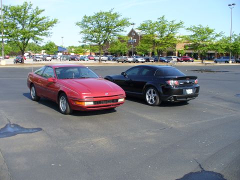 1991 Ford Probe - Two sports cars, a red 1991 Ford Probe and a black Mazda RX-8, are parked in a lot with a store in the background.