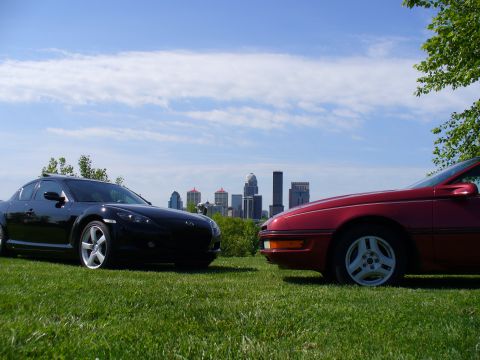 1991 Ford Probe - Two cars are parked on a grassy hill with a city skyline under a cloudy blue sky.