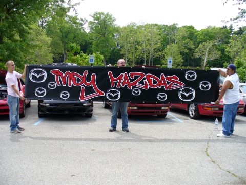 1991 Ford Probe - A group of people display a banner that says 'MDU MAZDAS' in front of multiple Mazda vehicles parked in a lot.