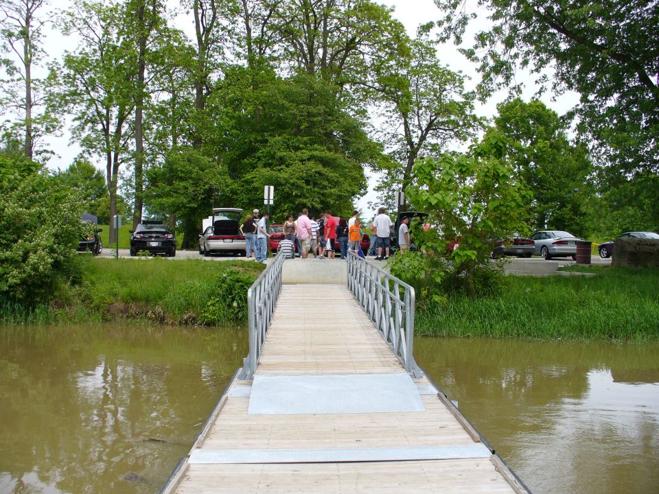 People gathered around cars parked near a river with a wooden walkway in the foreground