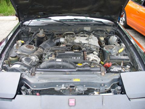 1991 Ford Probe - Engine bay view of a vehicle, with an orange car visible in the background. The engine is labeled 7M-GE.