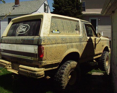 1989 Ford Bronco - This heavily mud-splattered Ford Bronco is viewed from the rear 3/4 angle, parked between two houses.