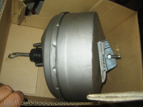 1990 Ford Bronco - silver vacuum power brake booster with pushrod sitting in an open cardboard box, with a parts label attached.