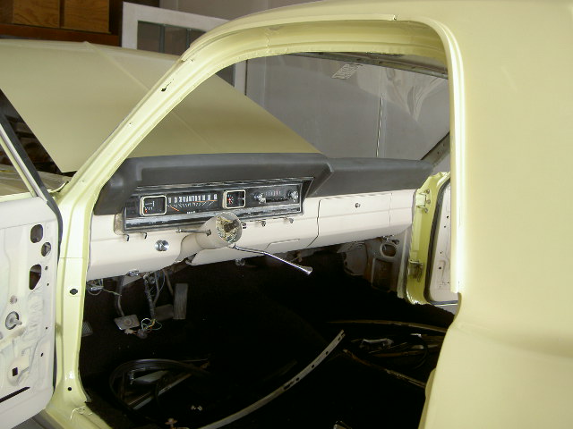 Interior shot of a yellow car, showing the dashboard, steering wheel, and pedals. The door is open