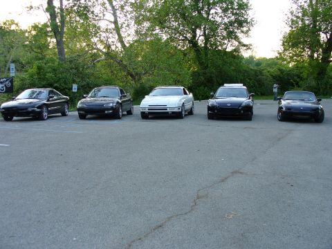 1991 Ford Probe - A line of five cars, including a black 1991 Ford Probe, are parked in an asphalt lot with trees in the background.
