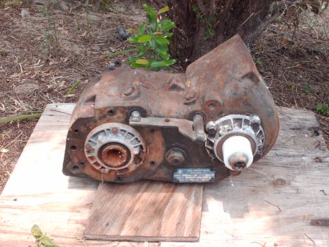 1982 Ford F-100 - A heavily rusted transfer case, possibly from a truck, sits on wooden boards with vegetation in the background.