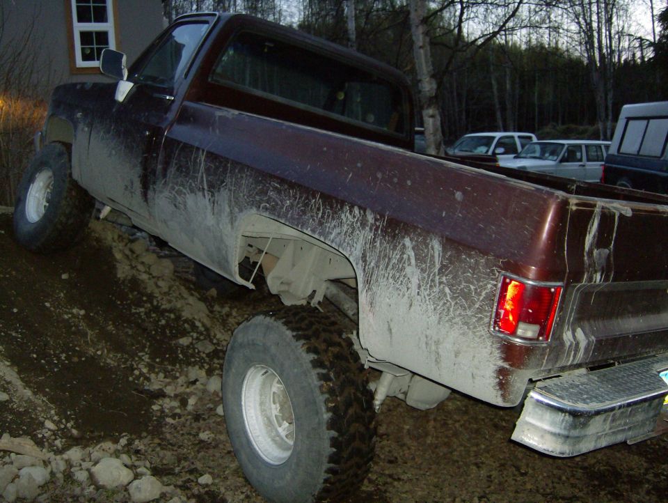 Mud-covered 1982 Chevrolet C/K Pickup truck angled on a dirt slope, with other vehicles visible in the background
