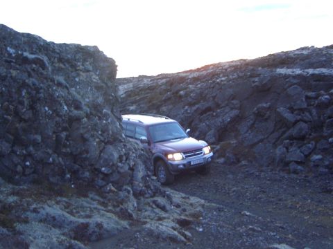 1974 Ford Bronco - A red SUV drives through a rocky, uneven terrain, its headlights illuminating the path ahead.