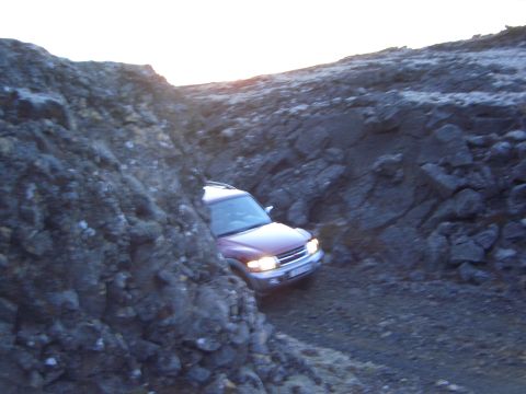1974 Ford Bronco - A red SUV carefully drives through a narrow, rocky canyon, with the sun setting in the background.