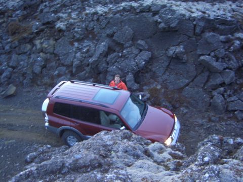 1974 Ford Bronco - A red SUV is shown driving on a challenging, rocky terrain, with a person observing from behind.