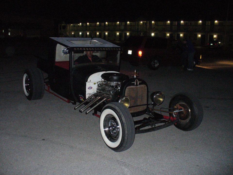 Black hot rod with exposed engine, white wall tires, and a custom roof, viewed from the front-right