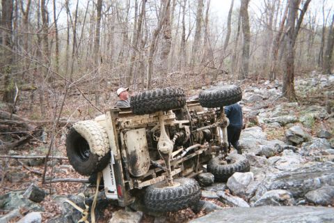 1969 Ford Bronco - An off-road vehicle has rolled onto its side in a challenging, rocky terrain with trees all around.