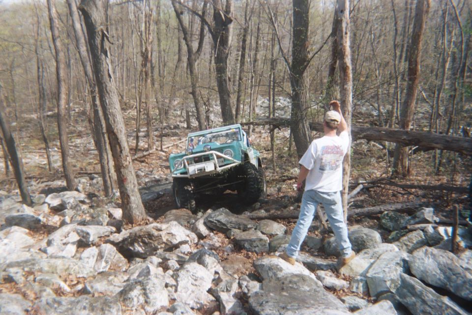 1969 Ford Bronco with custom bumper and roll cage driving over rocks in a wooded area