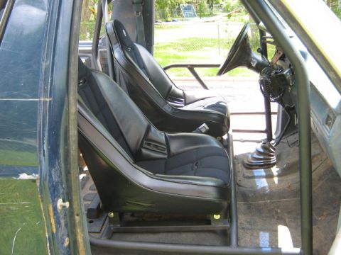 1986 Ford Bronco - The interior of a vehicle shows two bucket seats and a roll cage, with a view of the outdoors through the open door.