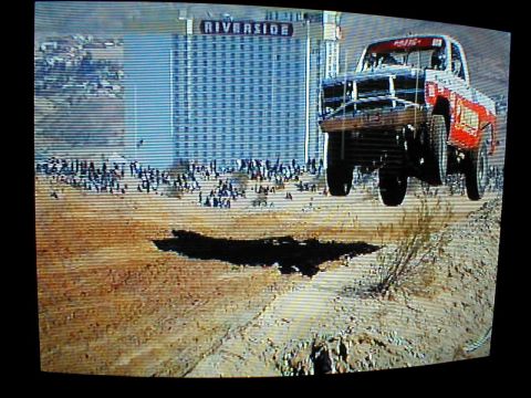 1996 Ford Bronco - A trophy truck soars over a dirt jump with a large hotel in the background.