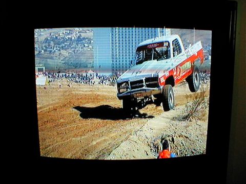 1996 Ford Bronco - A modified truck jumps during an off-road race with spectators and buildings in the background.