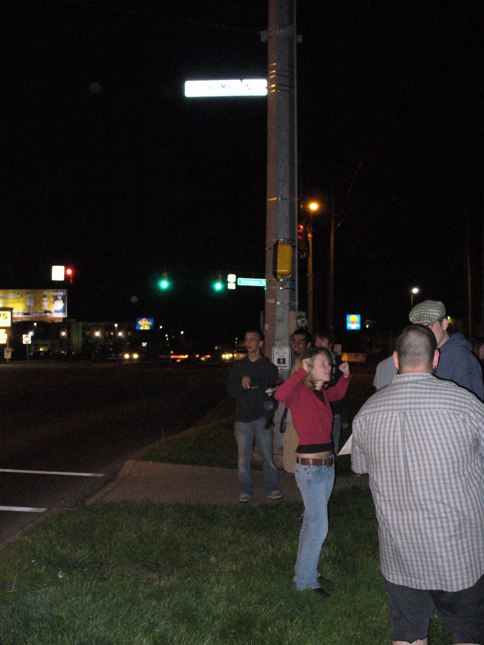 People stand on a corner at night near a street sign and traffic lights