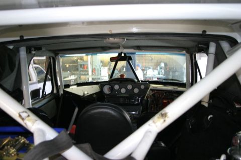 1996 Ford Bronco - Interior view of a race-prepped vehicle shot from the rear, showing a white tubular roll cage, a stripped dashboard with a cluster of round…
