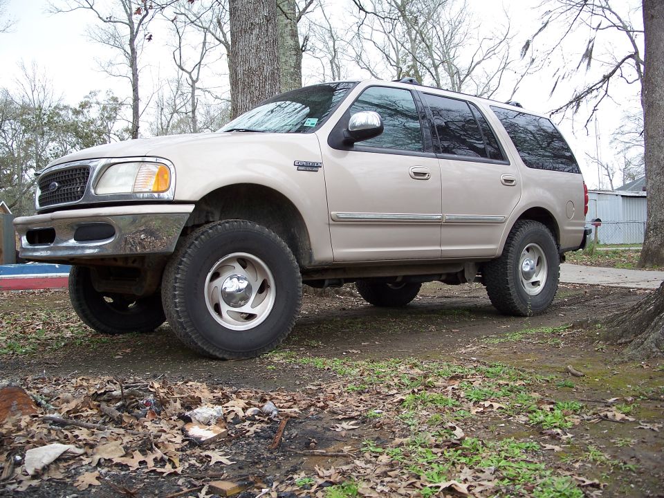 Tan Ford Expedition SUV with all-terrain tires parked on leafy dirt yard, front three-quarter view
