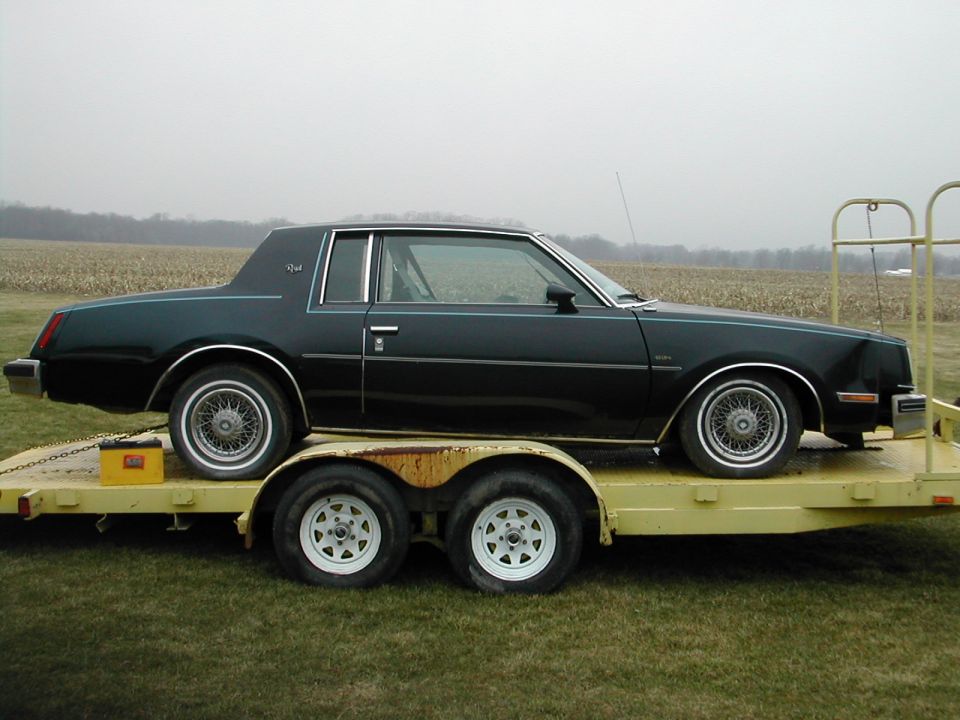 Black 1980s two-door coupe with wire wheels sitting on a yellow flatbed trailer in a field