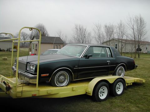 1998 Ford Crown Victoria - A black coupe with distinctive wire wheels is being transported on a yellow trailer.