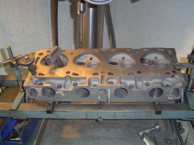 Bare cylinder head mounted on a machine shop fixture under a drill press for valve seat work