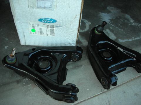 1998 Ford Crown Victoria - Two black-painted lower control arms with ball joints next to a Ford-branded parts box on a concrete floor.