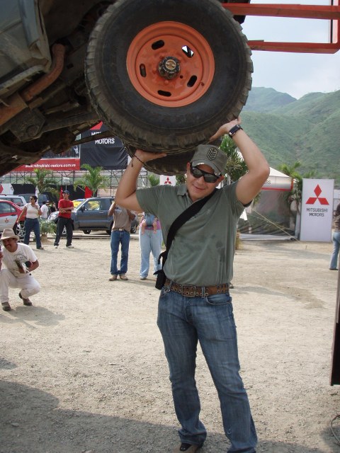 1995 Ford F-150 - A man lifts a large tire above his head in an outdoor setting with other people and vehicles visible in the background.