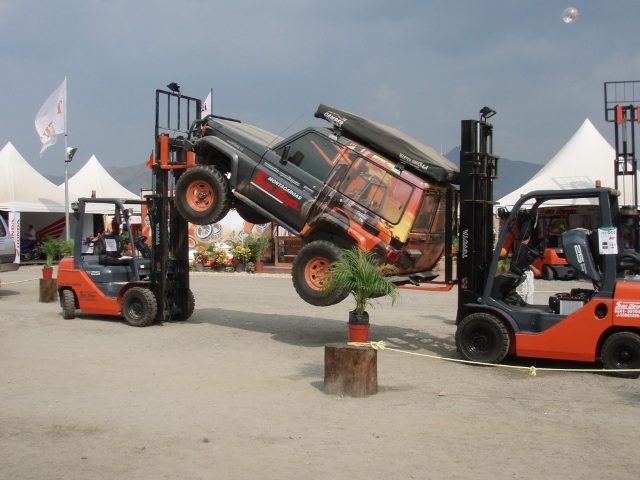 Toyota Land Cruiser with a rooftop tent being lifted by two forklifts, one on each side. Another forklift is visible in the background