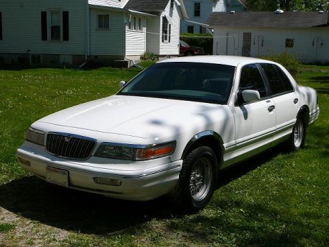 1997 Mercury Grand Marquis - Driver-side front three-quarter view of a white full-size Mercury four-door sedan on grass beside a white house, showing the vertical-bar g…